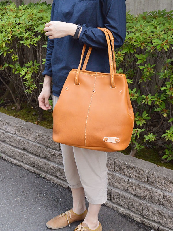 棕色皮革托特包，設有可調式提把，包身有白色縫線裝飾，一側附有品牌標籤。