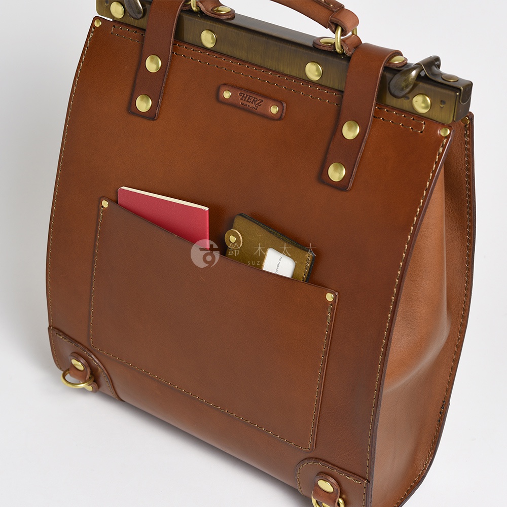 職人手作皮革復古口金後背包，採用棕色皮革材質，箱型設計，正面有兩個外袋，分別放入紅色護照和卡片夾。包包頂部有金屬提把和金屬口金鎖扣，搭配鉚釘和皮革綁帶。