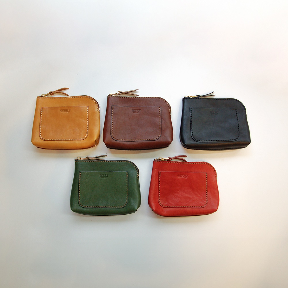 五款職人手作皮革小物收納包，顏色包含：淺棕色、深棕色、黑色、綠色、紅色。包款皆為方形，表面有縫線裝飾，並設有一個方形外袋，上方配有拉鍊開合。