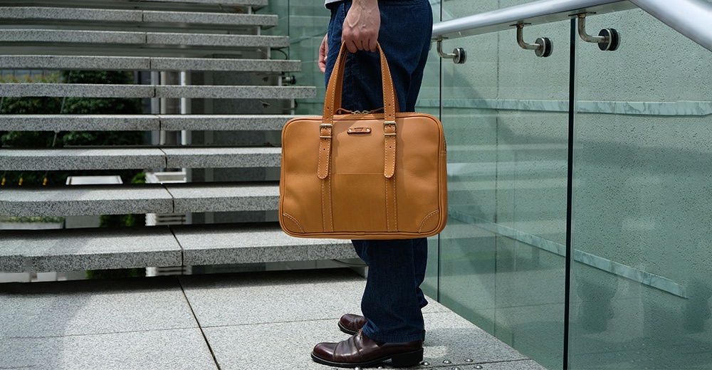 一個棕褐色皮革製的公事包，配有兩個提把和可調節的肩帶，包身有縫線細節和一個前袋。