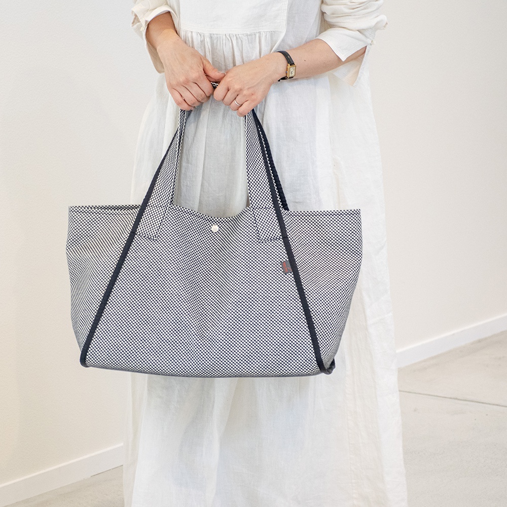 一位女士身穿白色長洋裝，雙手提著一個藍白格紋的三河木綿刺子織大容量托特包，包邊飾有深藍色滾邊。