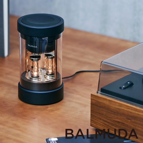 The Speaker 無線揚聲器，黑色圓形頂部帶有網格狀揚聲器，下方為透明玻璃圓筒，內部可見兩個發光的真空管，底部為黑色圓盤底座。