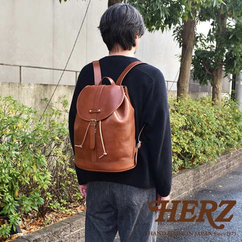 一位男士身穿黑色上衣、灰色長褲，背著一個棕色皮革單扣束口後背包。後背包採用光滑皮革製作，具有束口設計和金屬扣環，肩帶為皮革材質，整體風格簡約。