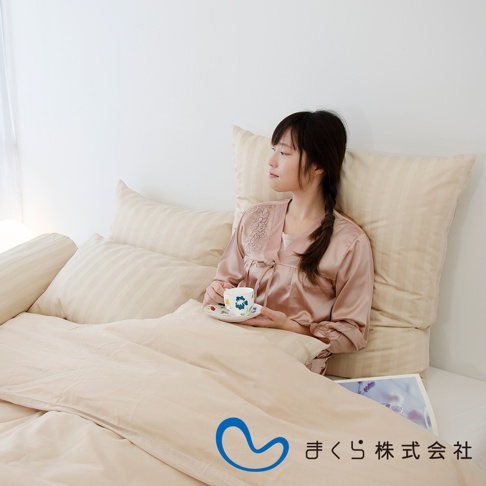 一位女性斜躺在床上，身穿絲質睡衣，手持印花杯碟。周圍是多個淺米色條紋的枕頭和床單，營造出舒適的居家氛圍。