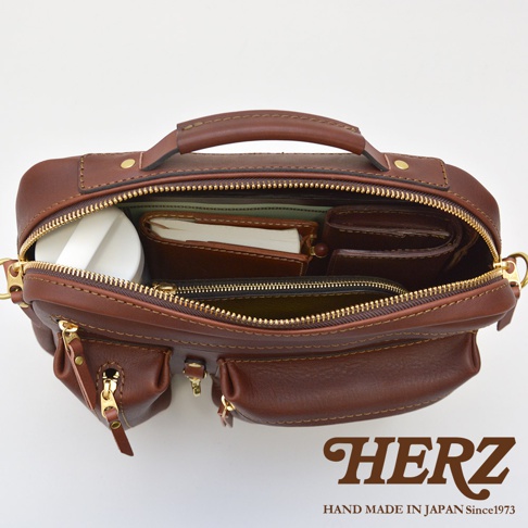 職人手作的棕色皮革兩用單肩包，內部可見多個隔層，裝有筆記本、錢包和圓形容器。包款設有拉鍊開合、頂部提把和外部小口袋，材質為皮革，金色拉鍊。