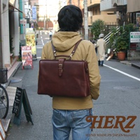 職人手作的咖啡色皮革軟式杜勒斯包，帶有金色金屬釦環和提把，肩背帶連接至背包。