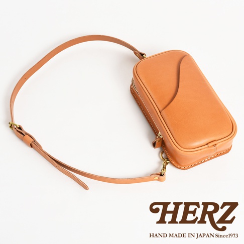 日本HERZ手工皮革製品，一個棕褐色的斜背包，配有長長的皮革肩帶，包身上有精緻的縫線和一個曲線設計，底部有HERZ的品牌標誌和「HAND MADE IN JAPAN Since1973」的字樣。
