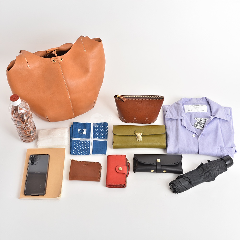 職人手作皮革球形肩掛托特包，搭配咖啡色拉鍊化妝包、軍綠色長夾、淺紫色襯衫、咖啡色零錢包、紅色卡夾、黑色小物包、摺疊傘，以及手機、水瓶和藍色圖案手帕。
