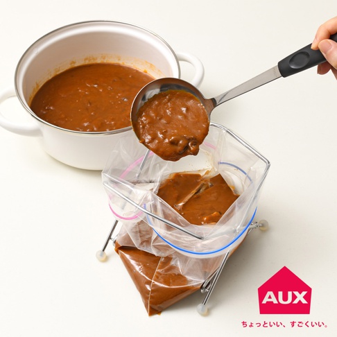 銀色金屬製成的折疊式萬用垃圾架，架上撐開兩個透明塑膠袋，袋內裝有棕色濃稠食物。一隻手持不鏽鋼湯勺，正將食物舀入其中一個塑膠袋。