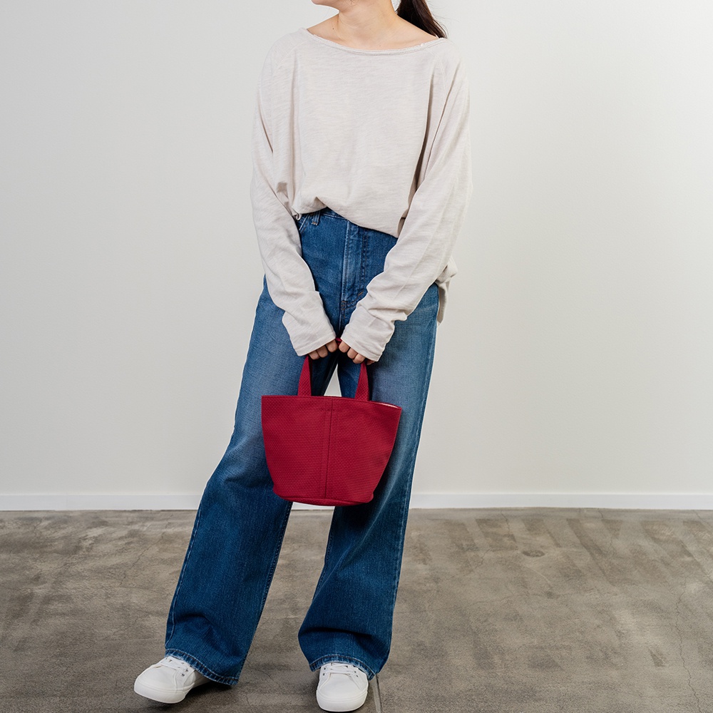 一位女士手持一個紅色圓底水桶包，包身由三河木綿刺子織布料製成，具有獨特的編織紋理。