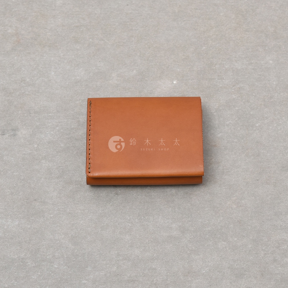一張從正上方俯拍的棕色皮革迷你短夾，簡約的雙折設計展現出義大利產皮革的細膩質感，左側邊緣有精緻的職人手縫線細節，短夾中央印有鈴木太太的品牌圖標與文字，整體呈現出極簡俐落的手作皮件風格。