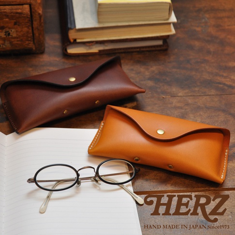 職人手作皮革眼鏡收納盒，一棕一橘，搭配復古眼鏡，背景為書籍與木桌。