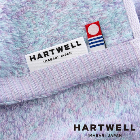 這款 Hartwell 今治毛巾呈現柔和的粉藍與粉紫色混色紋理，質感蓬鬆柔軟，特寫鏡頭下可清晰看見標籤上印有 Hartwell Imabari Japan 字樣，旁邊則附有一個帶有紅白圓形與藍色條紋圖案的精緻標籤。