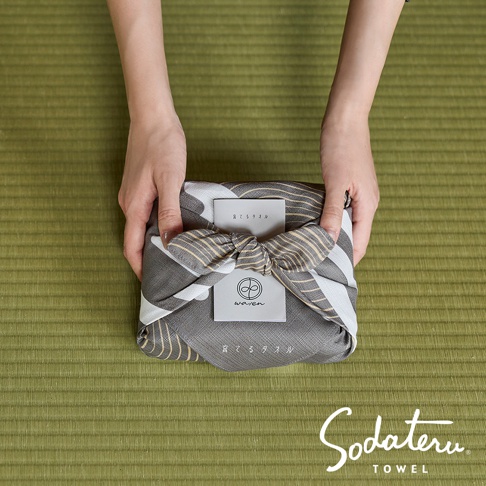 一雙手拿著一條印有灰白色條紋和金色線條的布包裹的禮物，布上繫著一個蝴蝶結，禮物正面有一張寫有日文文字和圓形圖案的卡片，下方為 Sodateru TOWEL 的品牌標誌。