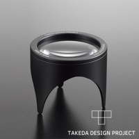 黑色鋁製閱讀放大鏡，鏡片清晰，鏡框周圍刻有「TAKEDA DESIGN PROJECT」及「Made in Japan」字樣，底部有三個彎曲的支腳。