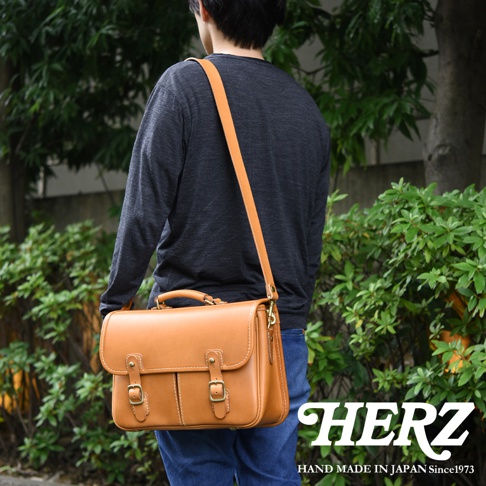 一款棕褐色皮革製成的兩用單肩包，包身設有兩個翻蓋和搭扣，頂部有手柄，搭配可拆卸肩帶，由一名男子肩背。