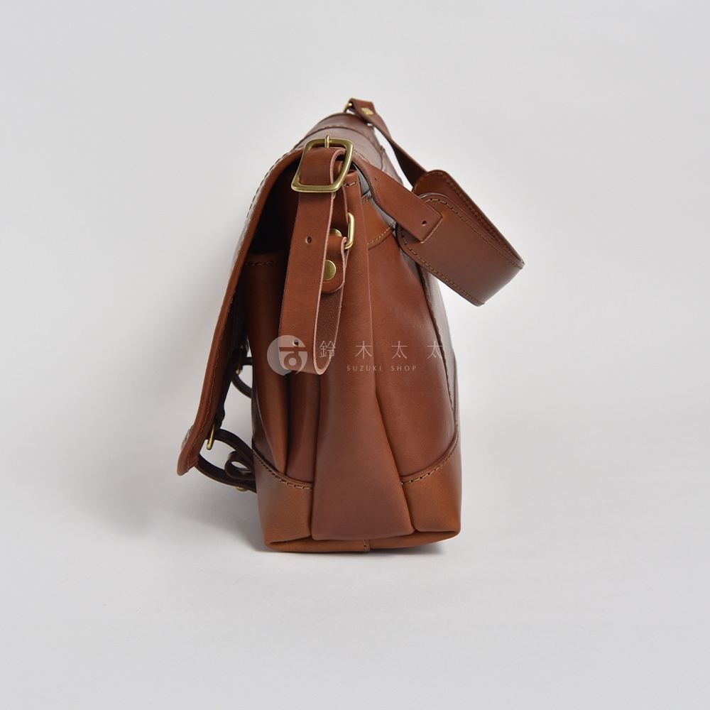 職人手作皮革復古郵差包 CM-100，這款包採用棕色皮革製成，側邊展示了其立體結構、可調節的肩帶和黃銅色金屬扣環，整體設計簡約而經典。