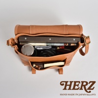 職人手作皮革復古郵差包，採用鞣製皮革製成，呈現自然焦糖色。包包內裝有書籍、保溫瓶、錢包等物品，側面有兩個小型外袋，其中一個裝有筆記本和零錢包。包包設計簡潔，適合日常攜帶。