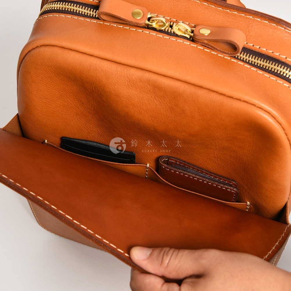 職人手作的棕色皮革後背包，展示其內部隔層，可放入卡片夾和錢包。包款採用黃銅色拉鍊和縫線細節。