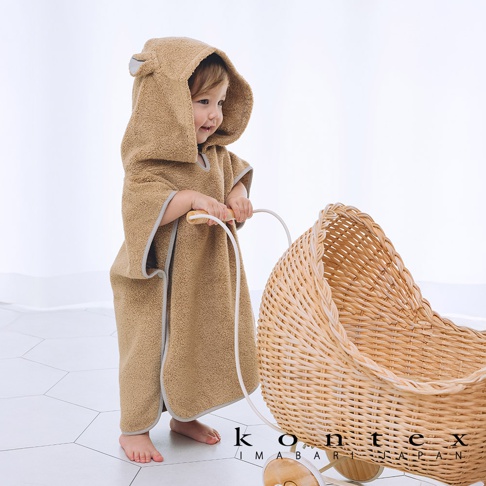 一位幼童身穿棕色毛絨斗篷式連帽浴巾，浴巾上有可愛的熊耳朵造型，浴巾邊緣有灰色滾邊。