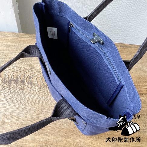 藍色帆布製成的水桶托特包，內部設有拉鍊隔層，搭配深色織帶提把，放置於木質表面。