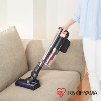 銀灰色金屬管身搭配黑色機身與透明集塵筒的無線手持吸塵器，吸頭為黑色，部分有藍色與紅色標示，正被一隻手持用在米色布質沙發上。