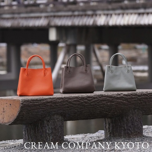 三款皮革手提托特包，分別為亮橙色、深棕色和淺灰色，皆配有提把，擺放在木質長凳上。