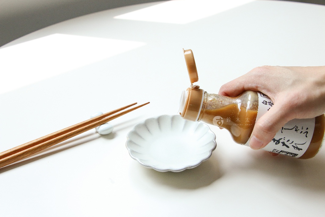 手持玻璃瓶裝的火鍋芝麻沾醬，將醬料倒入白色扇形小碟中，旁有一雙木質筷子架在白色筷架上。