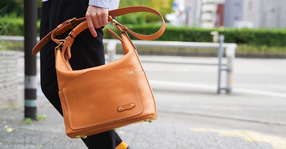 棕色厚實皮革單肩波士頓包，附有可調整長度的肩帶，包身有精緻縫線與品牌標誌。