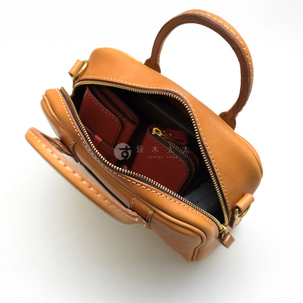 以鳥瞰視角拍攝的皮革迷你波士頓包，內部裝有紅色的皮夾和零錢包，包身為棕褐色，拉鍊為金色，左下方角落有「SUZUKI SHOP」字樣。
