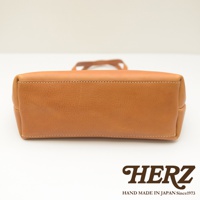 職人手作皮革托特包，呈現溫潤的淺棕色，材質為真皮，包體底部邊緣有細緻的白色縫線，整體設計簡約。