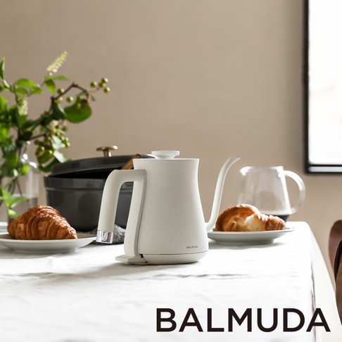 白色簡約手沖壺，配有直角手柄和細長壺嘴，置於白色桌布上，背景為深灰色鍋具、綠色植物與玻璃水壺，右側有可頌麵包。