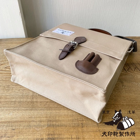 淺卡其色純棉帆布製成、帶有深棕色皮革飾邊的三用直式側背包，包蓋上有皮扣和皮革裝飾。