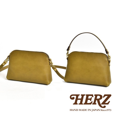 兩款淺棕色皮革製作的 HERZ 手工職人包並排展示，左側為配備長肩帶的肩背包樣式，右側則安裝了拱形手提帶，呈現可拆卸式的多功能設計，整體質感簡約且帶有經典手作皮革光澤。