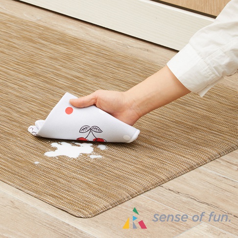 一隻手拿著印有紅色櫻桃圖案的白色擦拭墊，擦拭著表面帶有紋理的棕色廚房地墊上的白色液體。