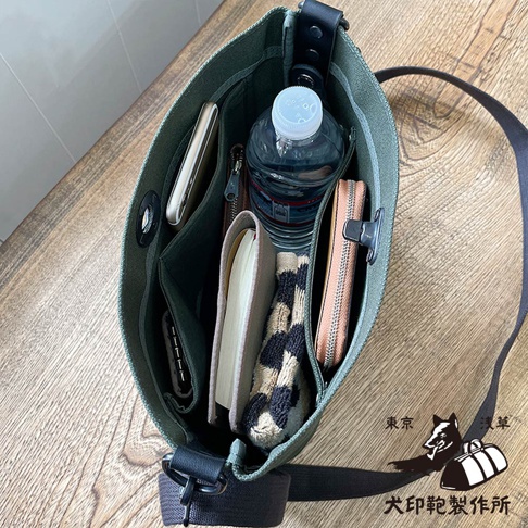 從上方視角拍攝，展示犬印鞄製作所的墨綠色帆布迷你側背包內部，可見手機、水瓶、皮夾、筆記本和一雙豹紋手套。