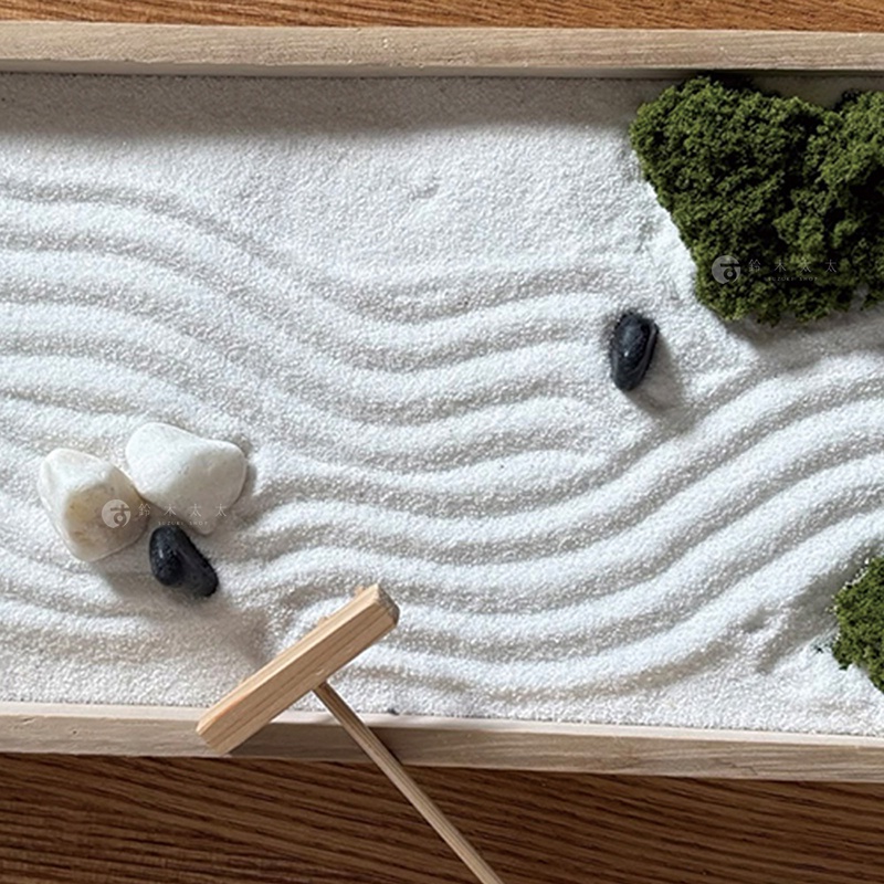 俯視視角下的一個長方形木製盒裝枯山水模型，白色的細沙上用耙梳出了平行的波浪紋理，角落處點綴著深綠色的青苔裝飾物，白沙之間錯落有致地擺放著幾顆黑色與白色的鵝卵石，畫面下方配有一支木製的迷你耙子。