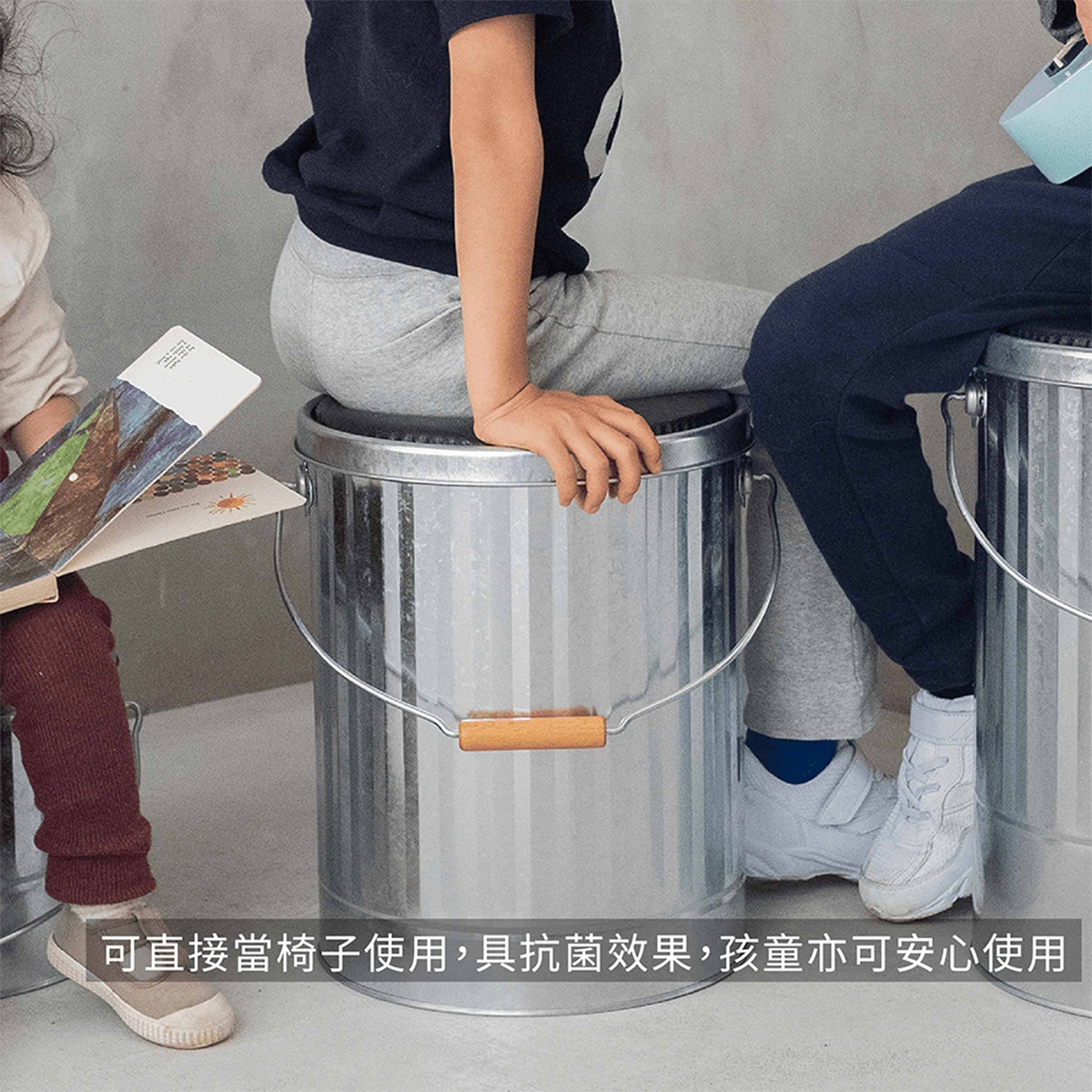 兩個孩子坐在銀色金屬儲物桶上，其中一個孩子正在閱讀一本圖畫書。
