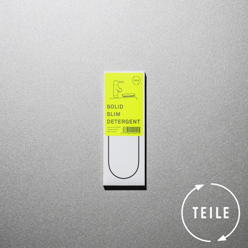 白色包裝盒頂部貼有螢光黃色標籤，印有黑色線條畫的廚房水龍頭和肥皂盤，下方標示「SOLID SLIM DETERGENT」。包裝盒下方有黑色U形輪廓。右下角有白色圓形標誌，內有箭頭和「TEILE」字樣。