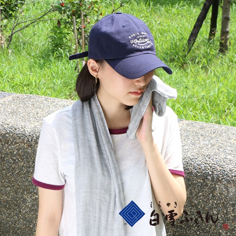 一位女性戴著深藍色棒球帽，身穿白色短袖上衣，頸部圍著一條淺灰色紗布長巾，並用長巾擦拭臉頰。