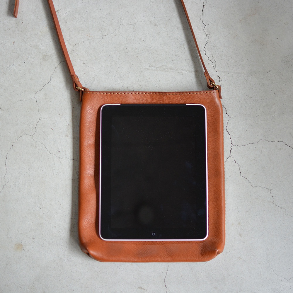 棕色皮革製成的肩背包，內置一台平板電腦，包包有肩帶，整體呈長方形，放在灰色水泥地面上。
