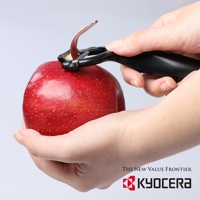 一隻手拿著紅蘋果，另一隻手使用黑色陶瓷削皮刀正在削皮，蘋果皮被捲起。