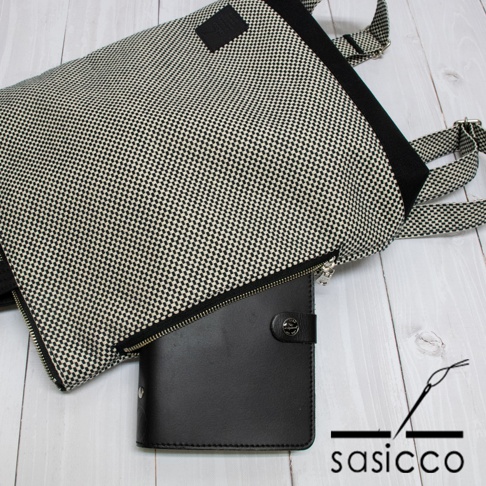 一款黑白格紋刺子織手提後背包，搭配黑色皮革記事本，背包有肩帶可調，材質為織布與皮革。