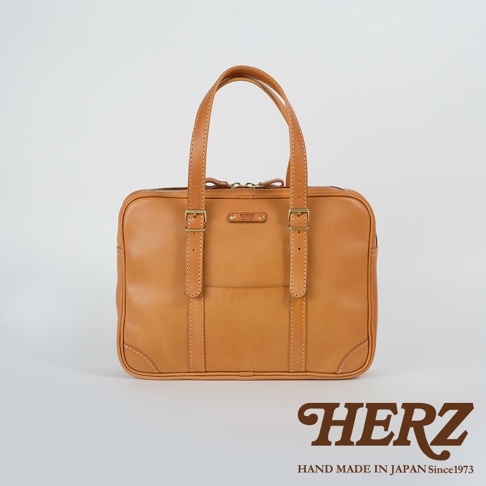 職人手作皮革肩背手提兩用公事包，呈現溫潤的淺棕色，主要材質為皮革。包款設計簡潔，有提把及可調節的肩背帶，前方有雙金屬扣帶裝飾，縫線細緻，整體風格經典。