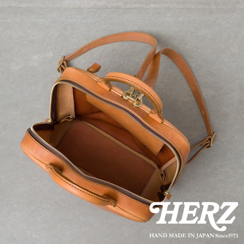 HERZ 職人手作皮革Organ商務後背包 M-10，採用天然皮革製成，呈現溫潤的焦糖棕色，包身為方形設計，開口處有金色金屬鎖扣，內部結構清晰，可見多個隔層，適合商務人士使用，展現細膩的工藝與質感。
