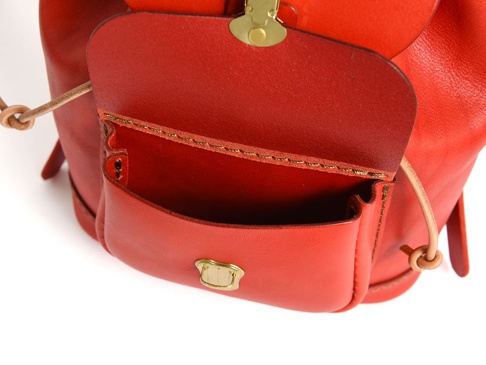 鮮紅色皮革迷你後背包，特寫其翻蓋前袋、抽繩與金色搭扣細節，展現職人手作質感。