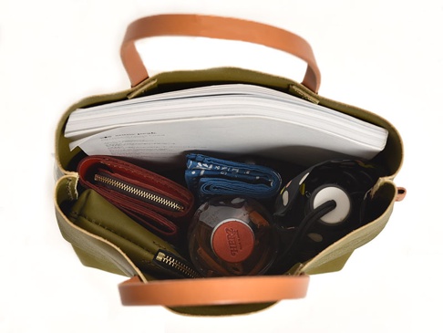 職人手作皮革直式軟皮托特包，橄欖綠色帆布包身，配有棕色皮革提把。包內裝有書籍、紅色錢包、藍色印花布、黑色圓點傘及一瓶琥珀色液體。