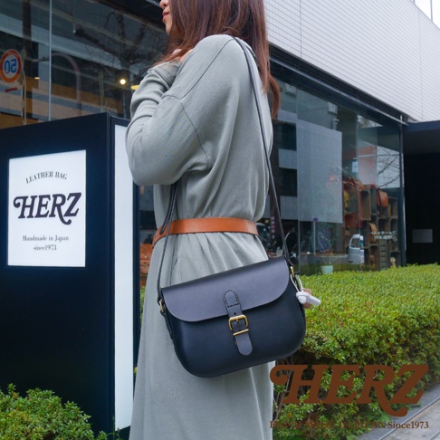 職人手作黑色皮革經典馬鞍包，配有金色釦環和棕色皮帶，由一位身穿灰色長洋裝的女士背著。