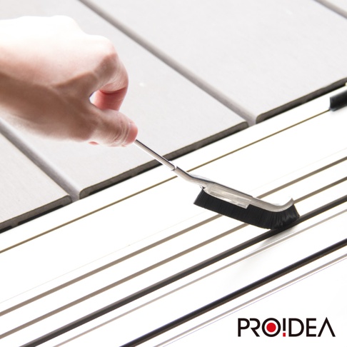 J型隙縫刷，擁有銀色金屬手柄，末端連接黑色密集刷毛，用於清潔淺灰色木紋地板的縫隙。