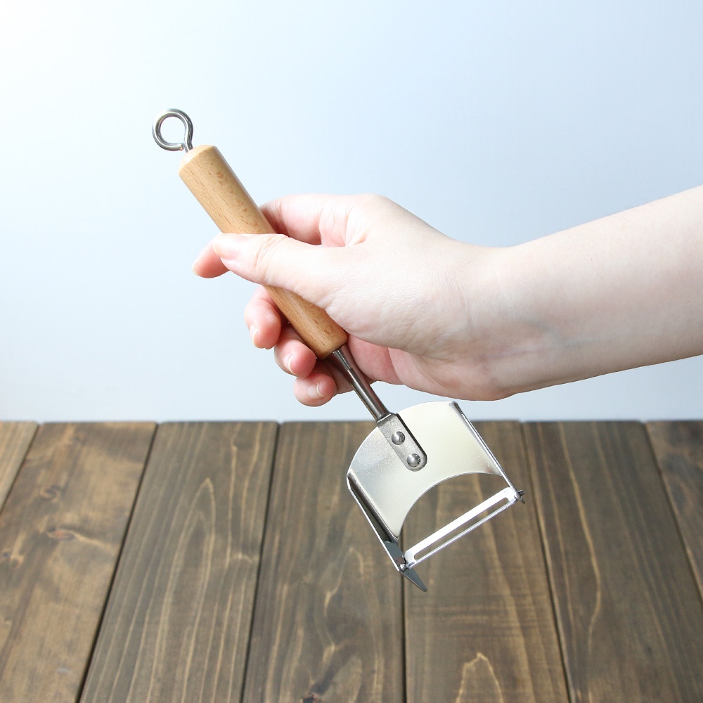 一隻手正握著 VEGET 不鏽鋼木柄削皮刀，刀柄為光滑的木頭材質，末端有金屬掛環。削皮刀的刀片部分為金屬製，呈弧形，設計簡潔。